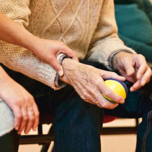 An elderly person receives support from a caregiver, holding hands indoors, showcasing compassion.